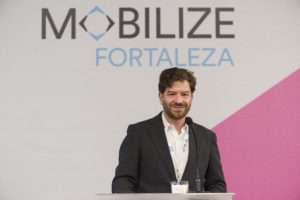 Man in jacket and white shirt stands before wall emblazoned with "MOBILIZE, Fortaleza"